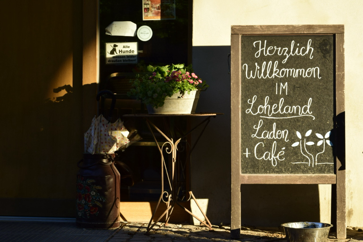 Willkommenstafel, Blumen und Schirmständer vor dem Eingang zum Café+Laden.