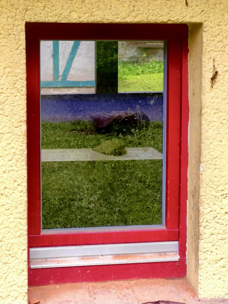 Blick durch zwei Fenster des Backhauses, mit Spiegelung und Dörrhaus dahinter.