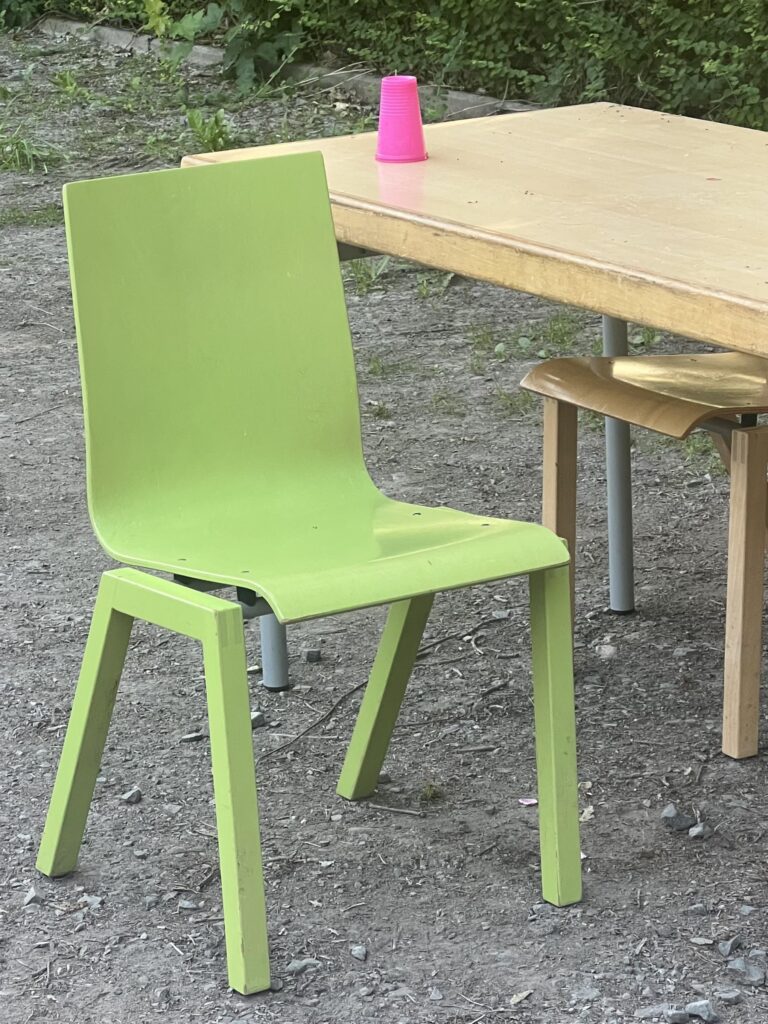 Reste des Getränkestandes von der Johanni-Feier: Schultisch mit grünem Stuhl auf dem großen Parkplatz. Ein pinker Becher steht auf dem Tisch.