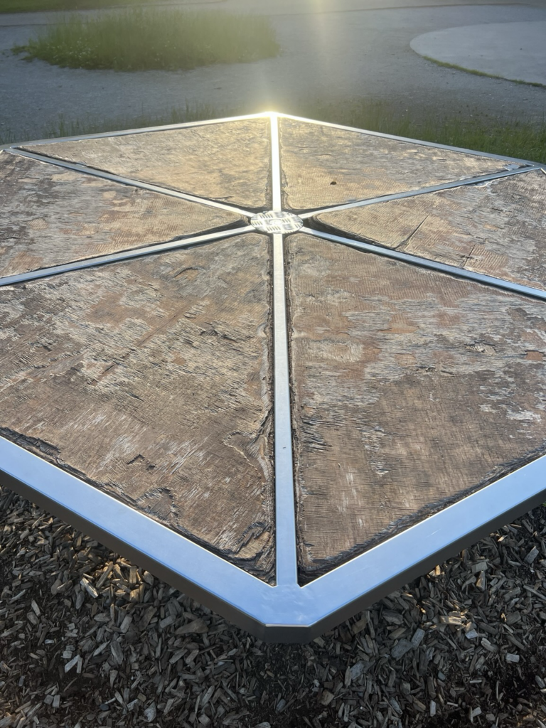 Sechseckiges Spielgerät auf dem Platz vor dem Haus für Spiel und Bewegung, die sechs Strahlen aus Metall zur Mitte reflektieren, die Abendsonne.