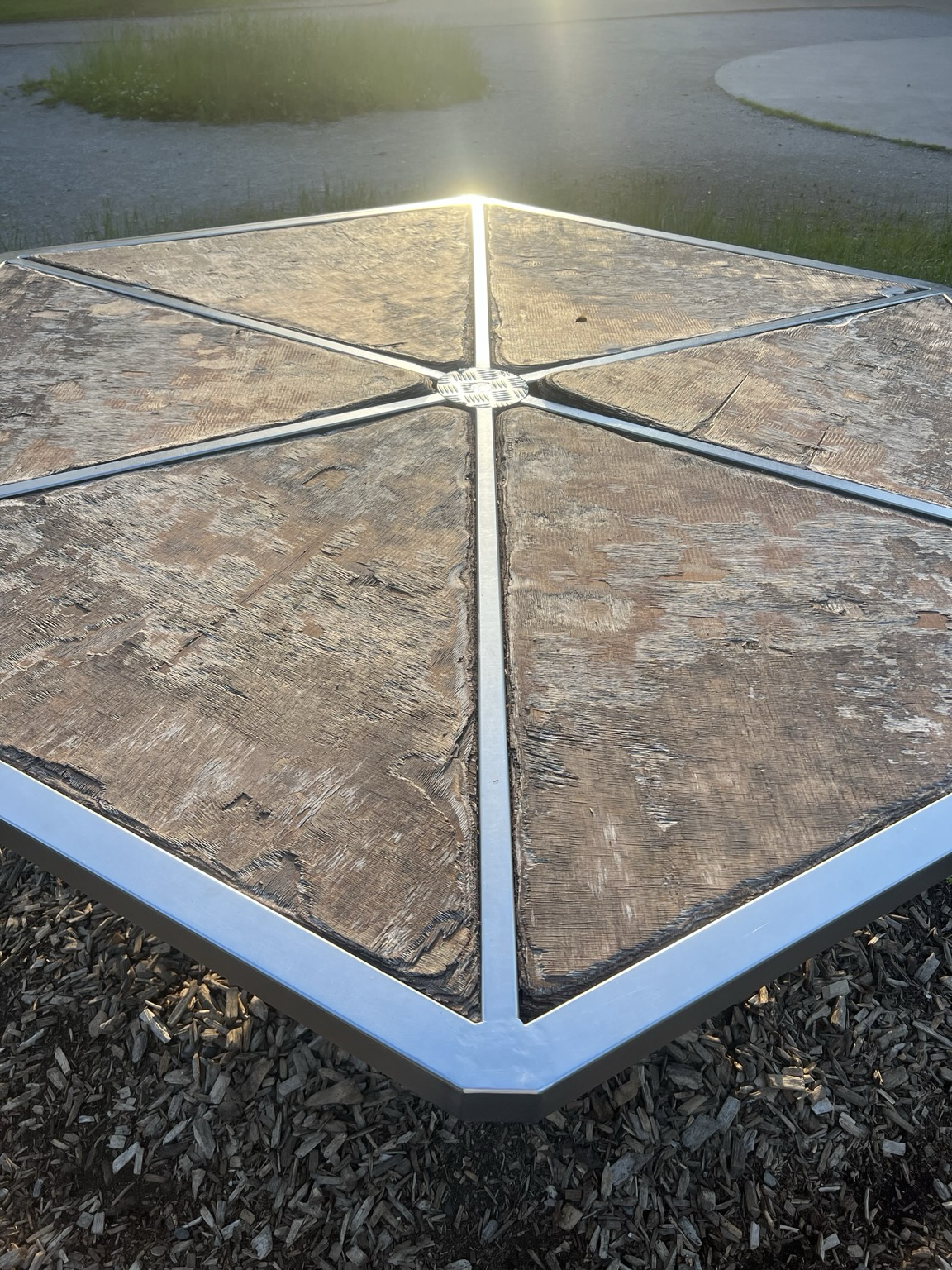 Sechseckiges Spielgerät auf dem Platz vor dem Haus für Spiel und Bewegung, die sechs Strahlen aus Metall zur Mitte reflektieren, die Abendsonne.