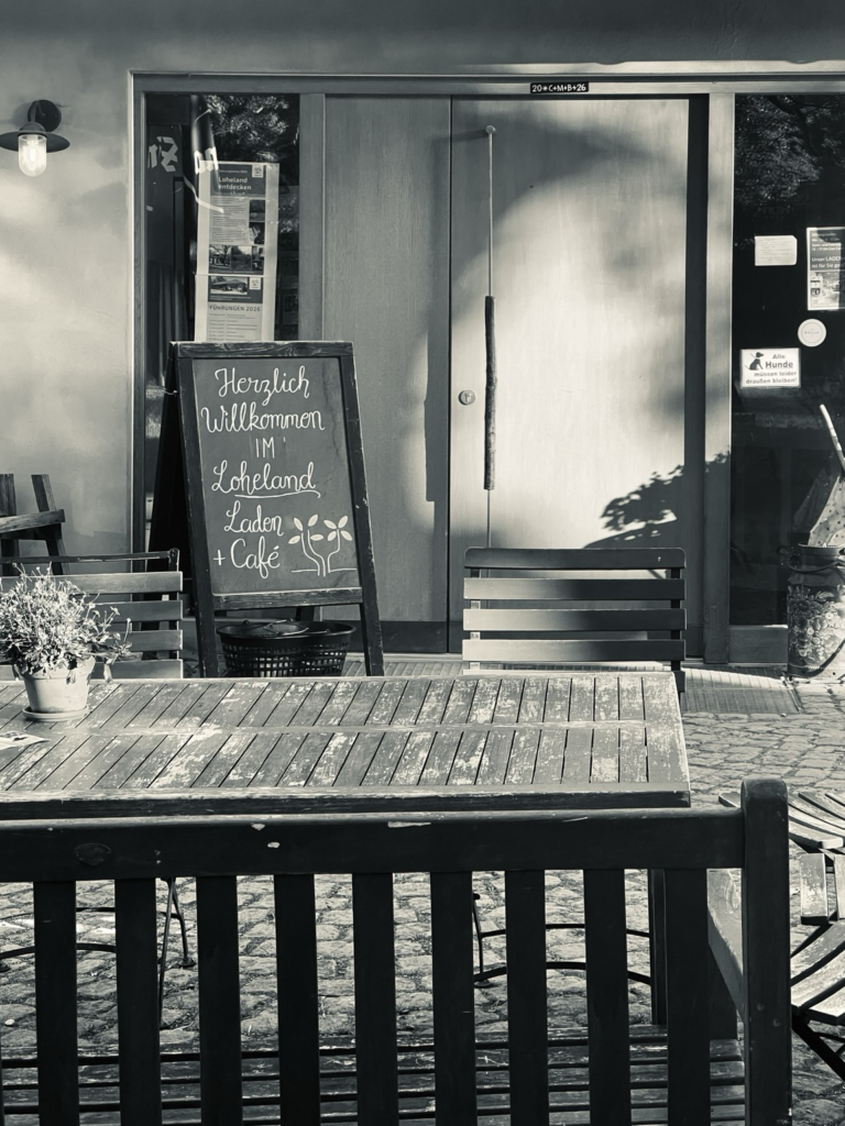 Eingang des Laden+Café in der Morgensonne, links von der Tür ein Willkommensschild, im Vordergrund ein Tisch mit Blumentopf und Stuhllehne des Stuhles dahinter.