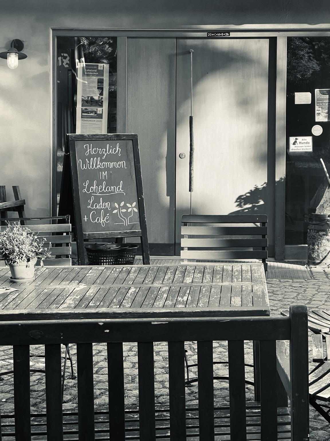 Eingang des Laden+Café in der Morgensonne, links von der Tür ein Willkommensschild, im Vordergrund ein Tisch mit Blumentopf und Stuhllehne des Stuhles dahinter.