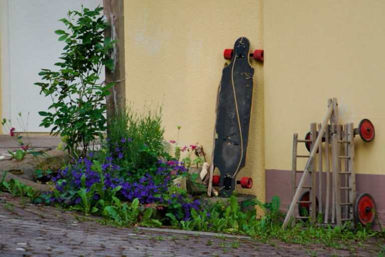 Longboard und Bollerwagen lehnen an der Wand des Mehrgenerationenhauses, davor Blumen, im linken Drittel ein unterer Teil der Haustür.